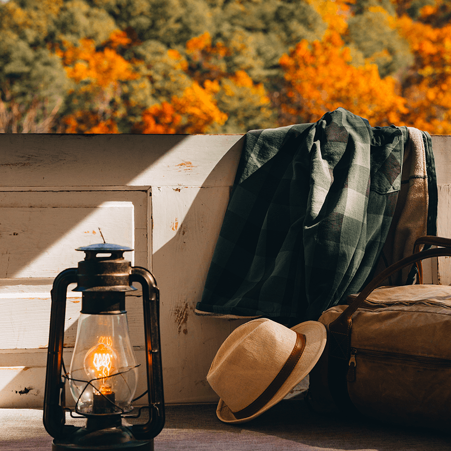 A vintage lantern, a plaid shirt, a straw hat, and a leather bag are arranged on a rustic surface with warm autumn foliage in the background.