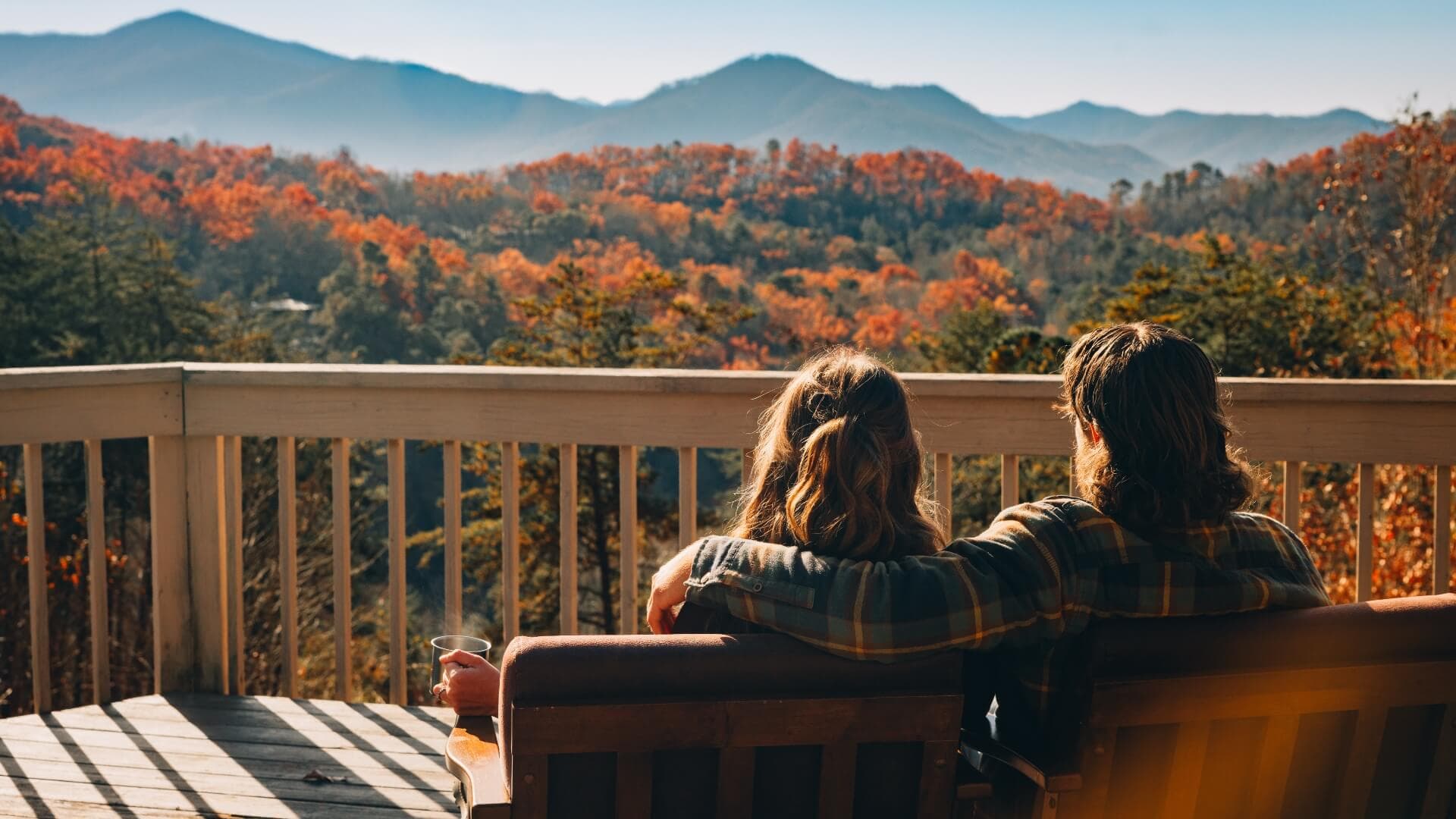 A couple enjoys a scenic view of mountains and fall foliage from a wooden deck.
