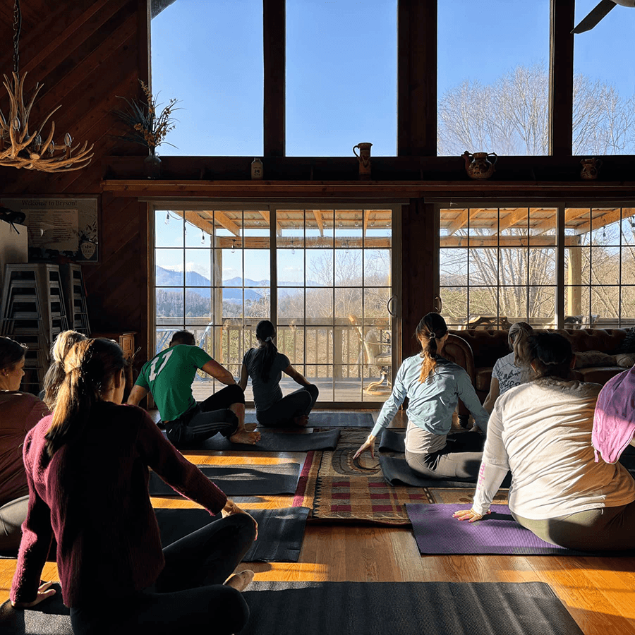 A group of people practices yoga indoors with large windows showcasing a scenic view of mountains and trees.