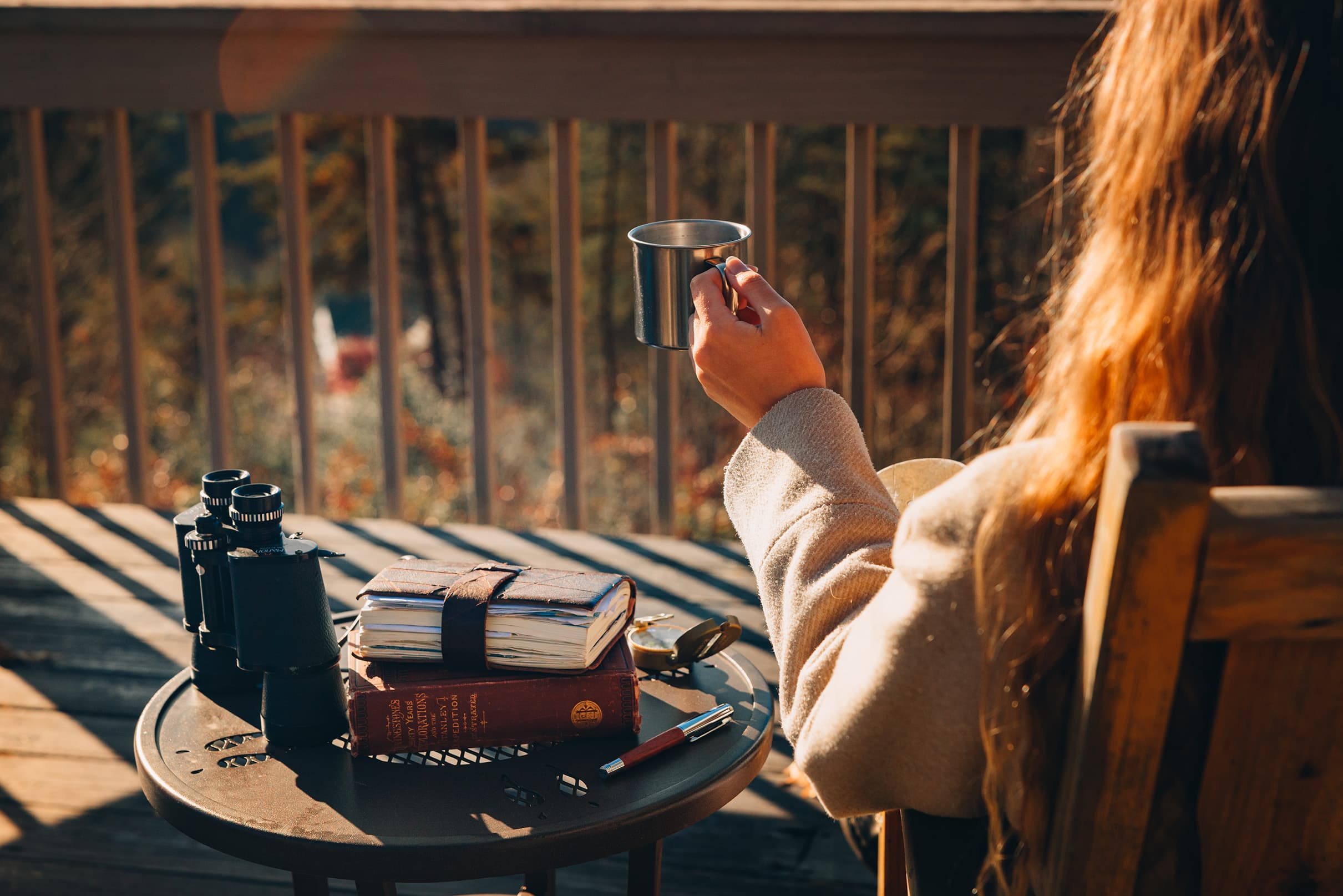 A person holds a metal cup while seated on a porch beside a table with binoculars and books.