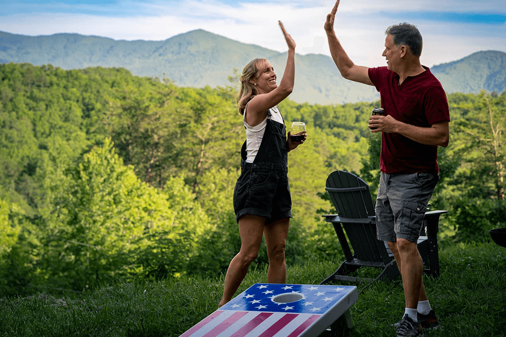 A woman and a man exchange high-fives outdoors against a backdrop of mountains, holding drinks and standing near a cornhole board.