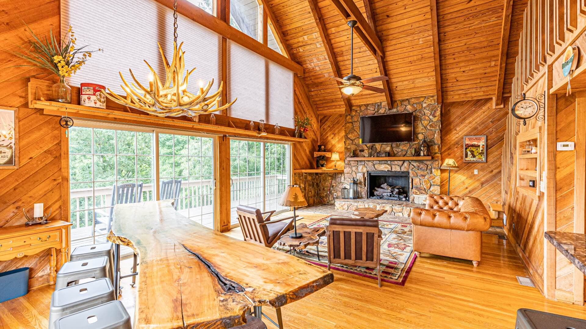 Cozy wooden interior of a cabin living room featuring a large table, a stone fireplace, and floor-to-ceiling windows.